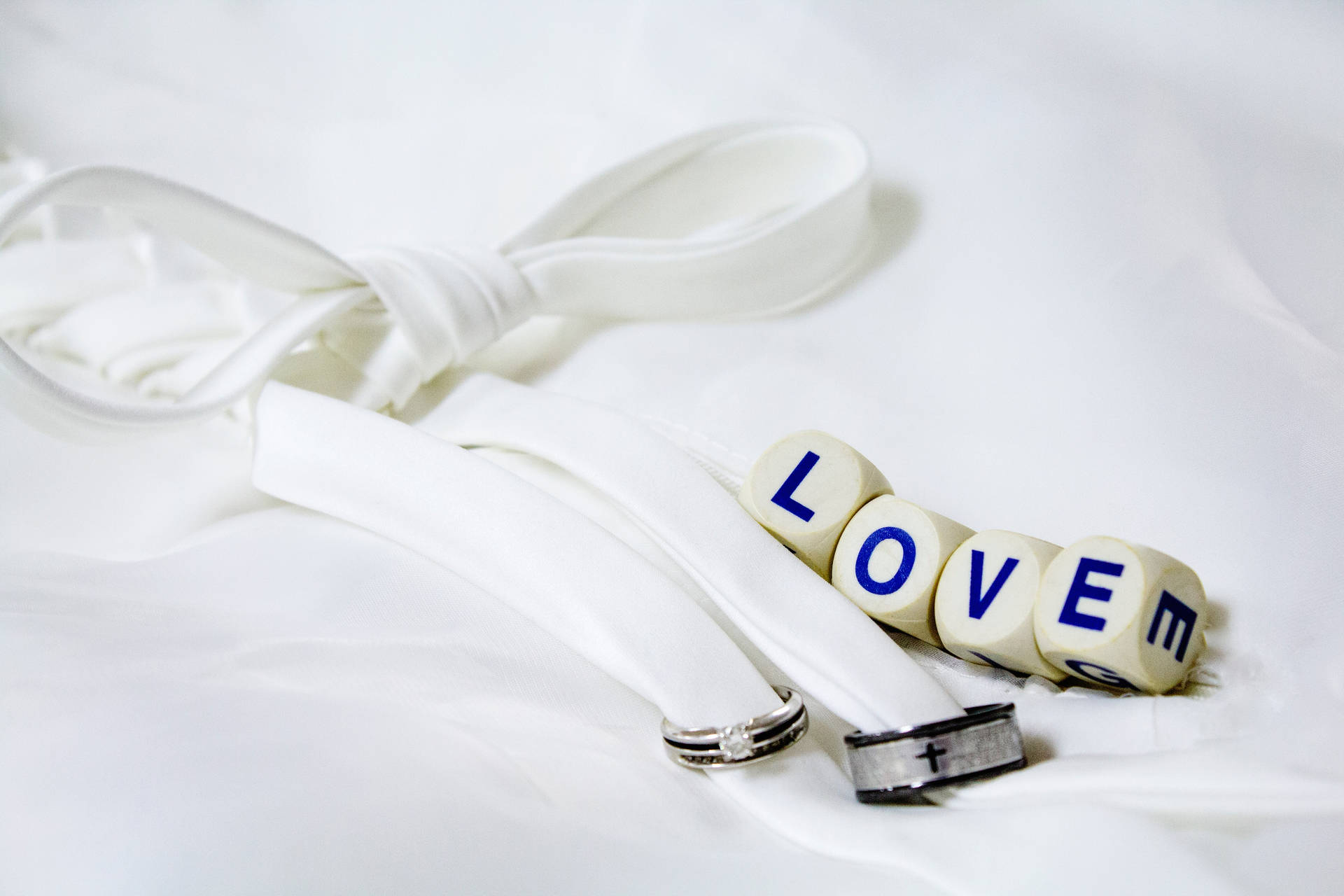 "Love" skrevet på fire hvide terninger ved siden af to sølv- og sort vielsesringe bundet med et hvidt bånd placeret på et hvidt lagen. — Kærlighed Wallpapers